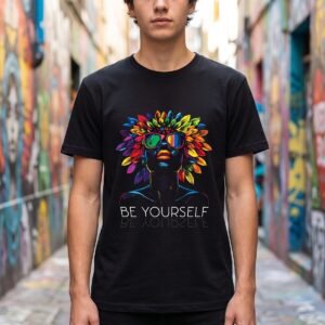 Person in a mural-covered alley wearing a black tee printed with a black profile in mirrored sunglasses under a crown of rainbow feathers, above Be Yourself